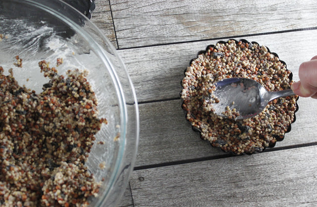 A hand uses a spoon to press a mixture of seeds into a small round tart tin, perfect for summer garden snacks. A glass bowl with more seed mixture sits beside it on a wooden surface.