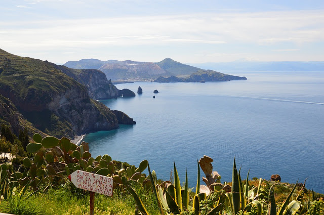 View of a rugged coastline with steep cliffs, clear blue sea, and distant islands—perfect for holidaying in Italy. Green vegetation and cacti are in the foreground, with a sign in Italian partially visible among the plants. The sky is clear and bright.