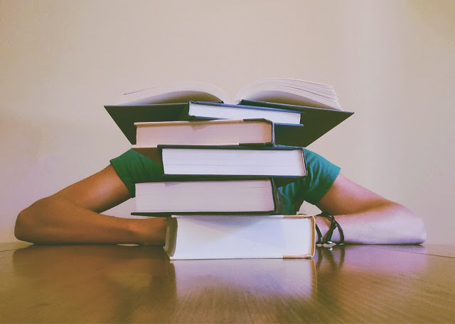 A person sits at a table with arms resting beside a tall stack of books, one open on top, obscuring their face. The scene captures the challenge of studying and hints at the need for revision tips to help your child study more effectively.