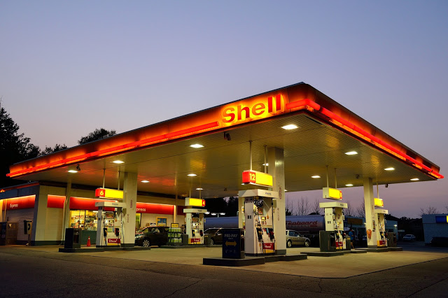 A Shell petrol station at dusk, brightly lit with red and yellow signage, showcases motoring history—multiple pumps and a convenience shop in the background reflect how times have changed, as several cars are parked nearby.