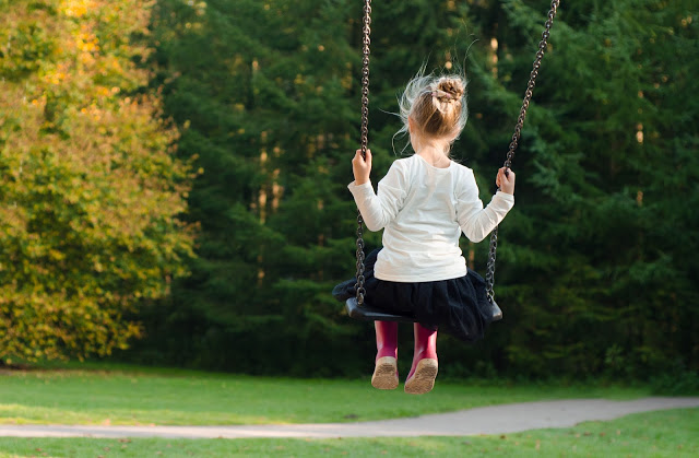 A shy child with blonde hair in a bun, wearing a white top, black skirt, and pink tights, sits on a swing facing away, surrounded by green trees and grass in a park—a moment that can help children boost confidence.