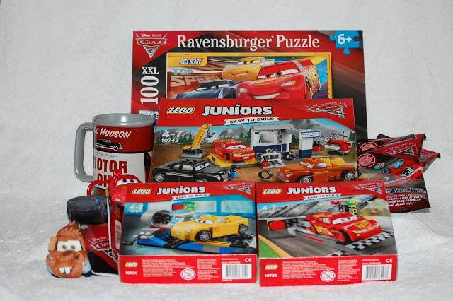 A collection of Cars 3 gifts, including a Ravensburger puzzle, LEGO Juniors sets, a silver mug, small soft toys, and two red-packaged surprises, all displayed on a white surface.