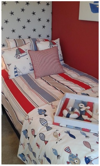 A single bed with nautical-themed bedding featuring boats and fish, red and blue stripes, and a striped cushion. The wall has black stars, and a tray with decor sits on the bed—perfect for families exploring child bed wetting solutions.