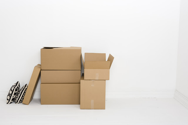 Several cardboard boxes, some open and some closed, are stacked against a white wall in a mostly empty room. Two patterned cushions—classic moving-in essentials—are leaning on the boxes to the left.