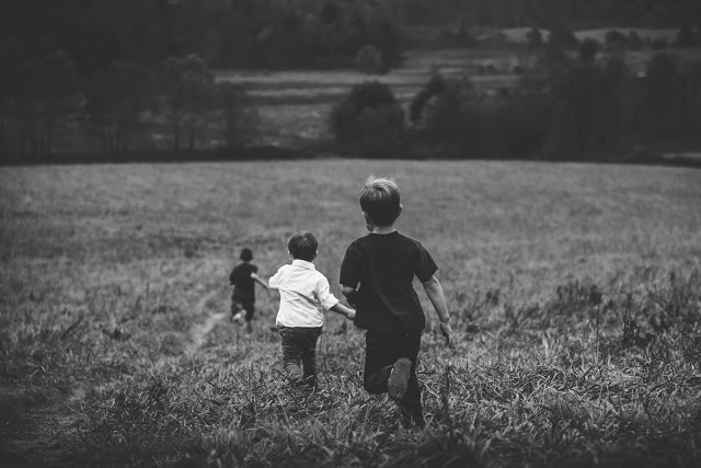 Three boys run through a grassy field towards a wooded area in the distance. The black and white image captures the children from behind as they move away from the camera.