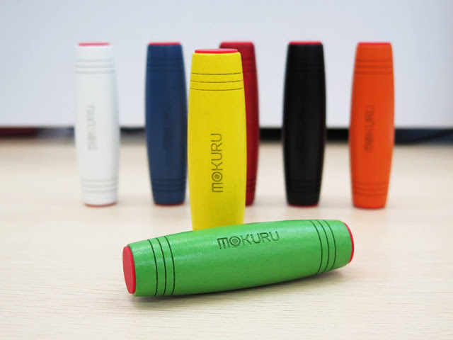 Seven colourful MOKURU fidget toys stand upright on a light surface, with a green one lying horizontally in the foreground—perfect for those looking to win at staying focused and entertained. Colours include red, orange, black, blue, white, yellow, and green.