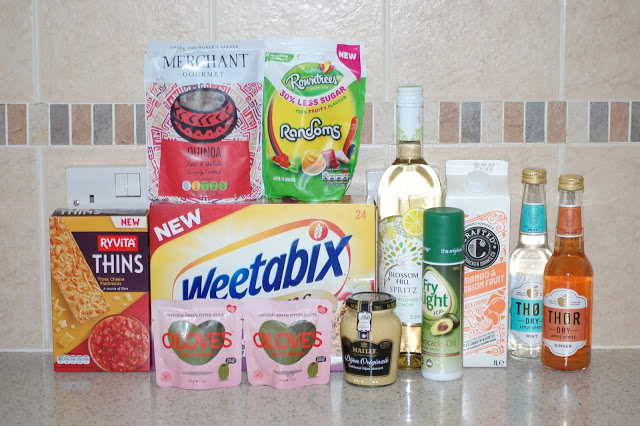 A selection of assorted food and drink items from the Summer Degustabox, including Weetabix, Ryvita Thins, olives, quinoa, Randoms sweets, white wine, fruit spritzers, Dijon mustard, and a tin of olive oil spray arranged on a kitchen worktop.