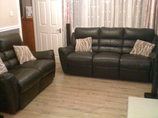 This inviting living room features two dark leather settees with patterned beige cushions on a light wood floor. Light-coloured curtains frame the window behind the settees, complemented by a white door to the left.