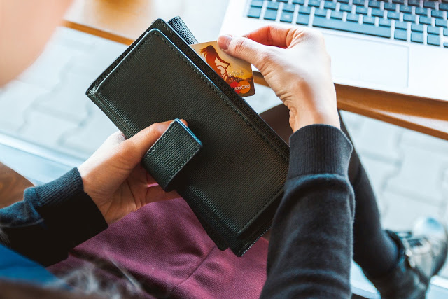 A person holding a black wallet and removing a Mastercard credit card, seated in front of a laptop on a table, considering ways to get money in an emergency.