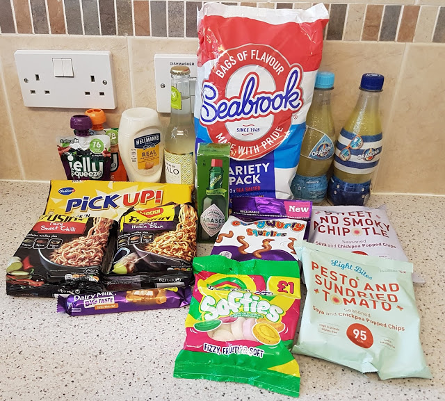 A variety of snacks and drinks from the Back To School Degustabox, including crisps, instant noodles, sweets, chocolate, flavoured water, and sauces, arranged on a kitchen worktop in front of a tiled splashback and electrical socket.