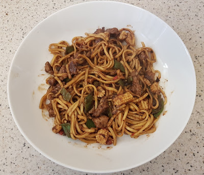 A white bowl filled with stir-fried noodles, pieces of meat, green vegetables, and a dark sauce sits on a speckled worktop—perfect for the #HealthyRedMeat Meal Challenge.