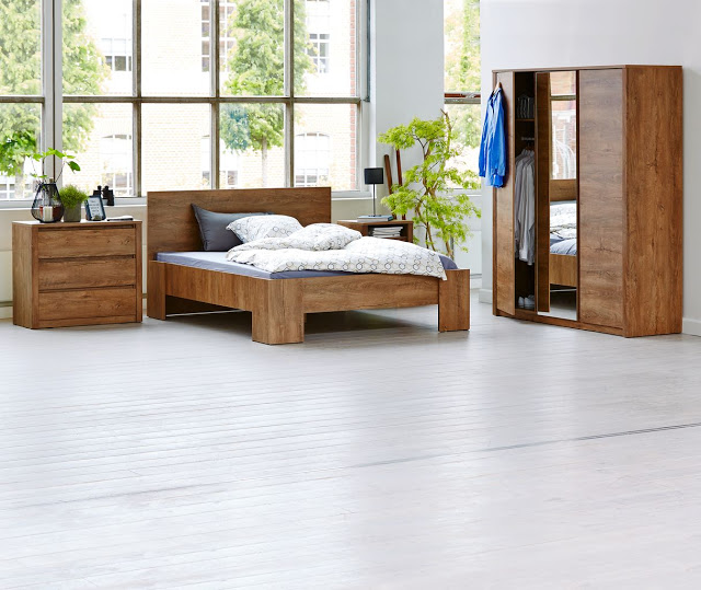A modern bedroom with large windows and JYSK wooden furniture creates a bright, minimalist space. Mirrored wardrobe doors, cosy Hygge touches like a plant, and clothes neatly hanging complete the inviting atmosphere.
