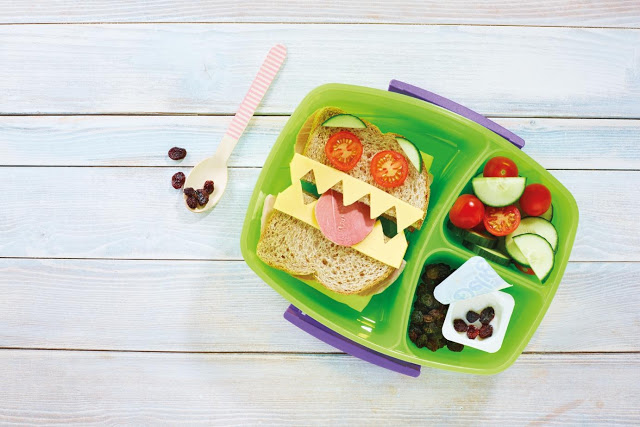 A green lunchbox with a sandwich decorated as a monster face using cheese, cucumber, tomato, and deli meat—one of many healthy lunchbox ideas—alongside cherry tomatoes, cucumber slices, raisins, and a yoghurt pot. A spoon and raisins sit on the wooden surface.