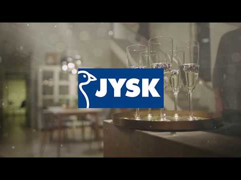 A tray with champagne glasses sits on a table in a warmly lit dining room, evoking true Hygge. The blue and white JYSK logo with its stylised bird is centred over the image.