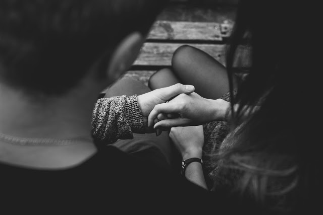 Black and white photo of two people sitting close together, viewed from above, gently holding hands on a wooden bench. Evoking intimacy, this image is perfect for articles on tips for couples who start living together. Only heads, arms, and hands are visible.