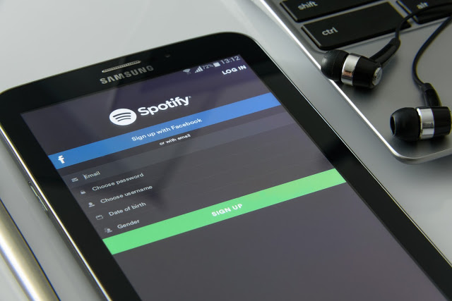A Samsung tablet displays the Spotify sign-up screen—one of the music must-haves—beside a laptop and black earphones on a desk. The Spotify screen shows fields for email, password, username, date of birth, and gender.
