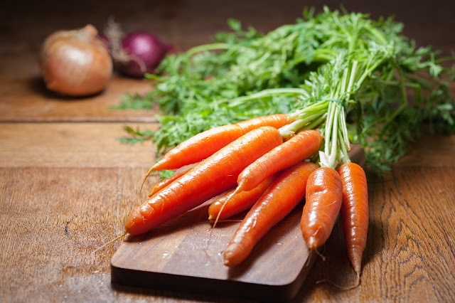 A bunch of fresh fruit and vegetables, including carrots with green tops, rests on a wooden chopping board, with onions in the blurred background on a wooden table.