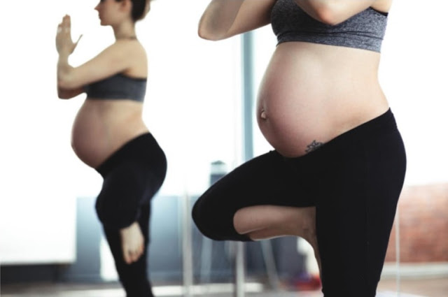 A pregnant woman, staying active during pregnancy, wears a sports bra and leggings as she practises a yoga pose indoors, balancing on one leg with her hands together, her reflection visible in the mirror behind her.
