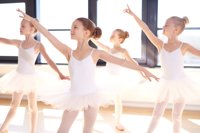 Four young ballet dancers wearing white tutus and leotards practise at a studio with large windows, arms gracefully extended and standing en pointe—showing some of the ways dance can benefit your child through discipline, poise, and confidence.