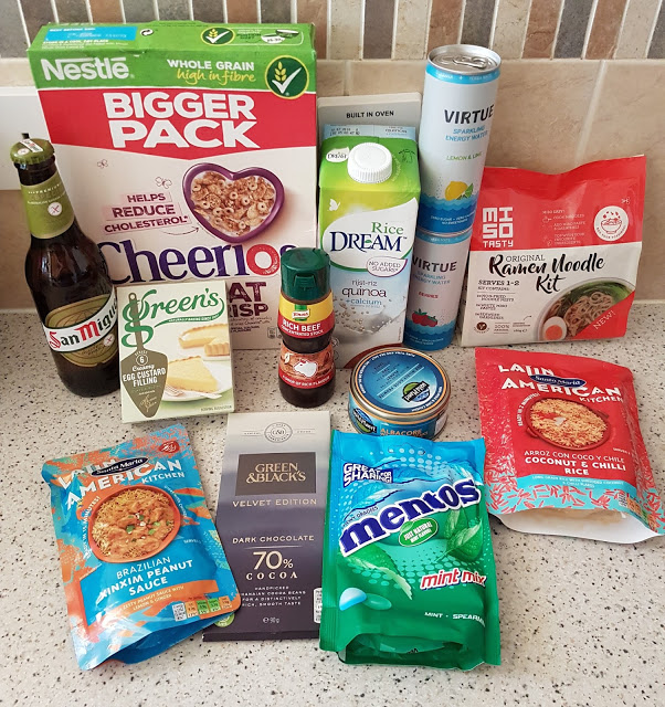 A selection of assorted groceries from an Autumn Degustabox sits on a kitchen worktop, including Cheerios cereal, rice milk, a noodle kit, tinned tuna, chilli con carne mix, mint Mentos, San Miguel beer, hot sauce, chocolate bars, and green tea.