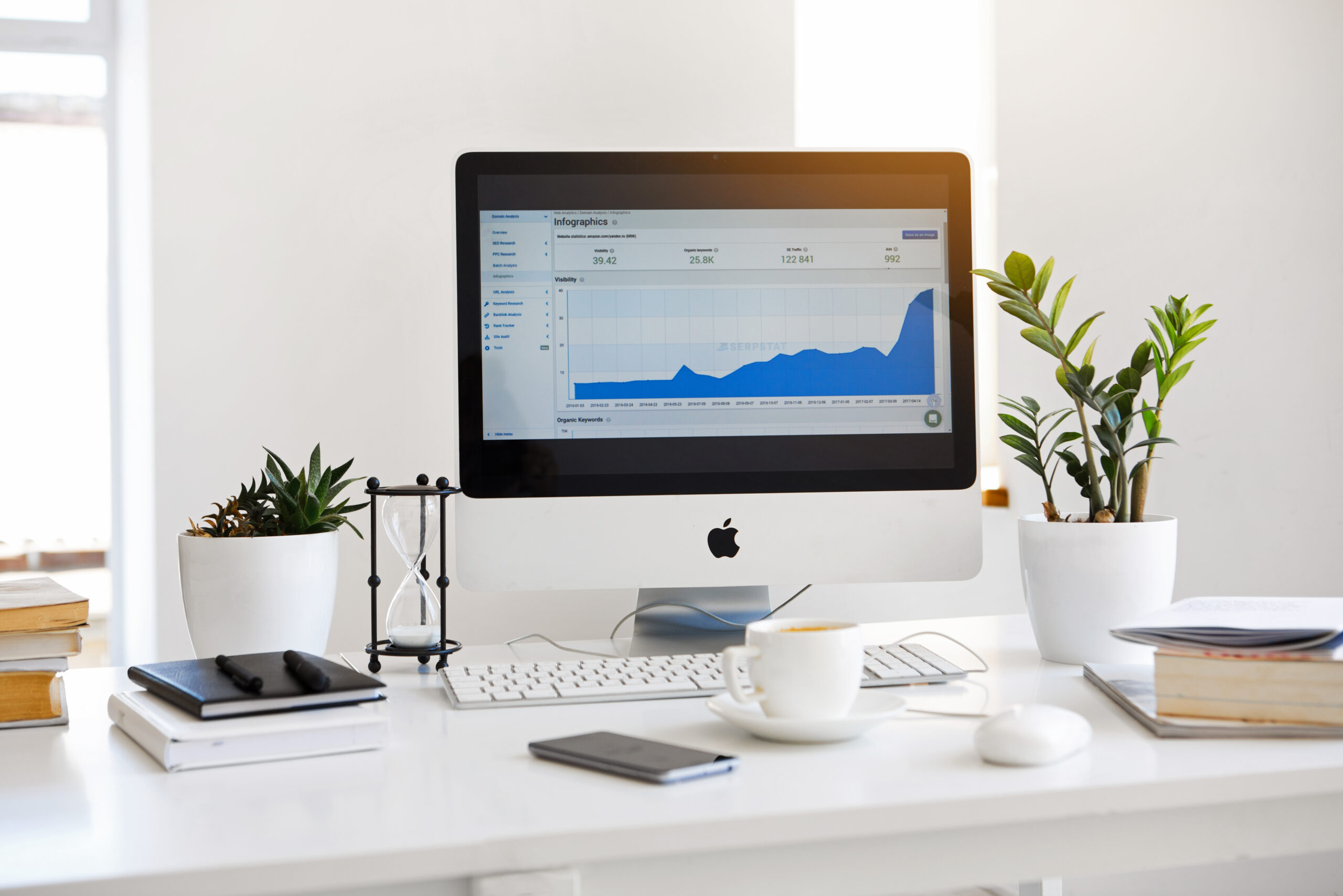 A modern desk with an Apple computer displaying a blue performance graph, surrounded by potted plants, notebooks, a cup of coffee, a mobile phone, and other office essentials in a bright, minimalistic workspace.