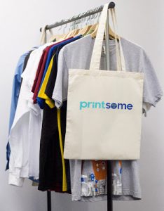 A beige personalised tote bag with the word "printsome" hangs on a clothing rail beside several colourful T-shirts and jumpers on wooden hangers against a plain background.