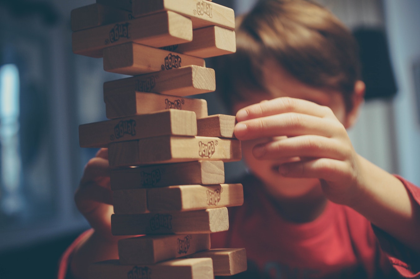 A child carefully removes a wooden block from a stacked Jenga tower, focusing intently on the game with both hands touching the blocks. The softly lit room creates a cosy atmosphere perfect for children to enjoy quiet play.