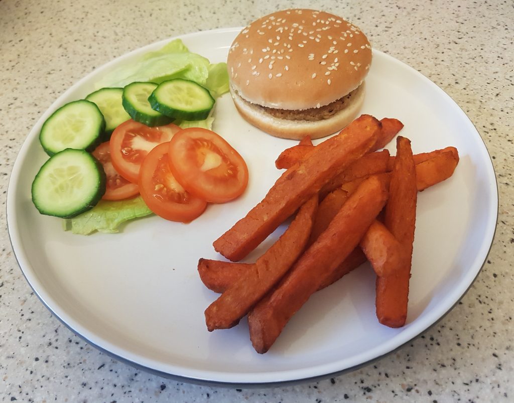 A plate with a Rustlers hamburger in a sesame seed bun, sweet potato chips, and a side salad of lettuce, cucumber, and tomato slices sits on a speckled worktop.