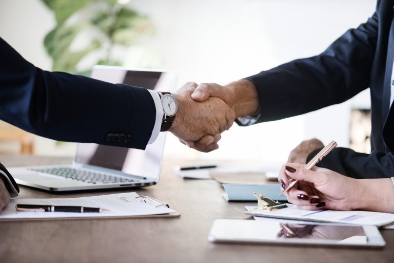 Two men shaking hands in an office with a lady writing notes in the foreground