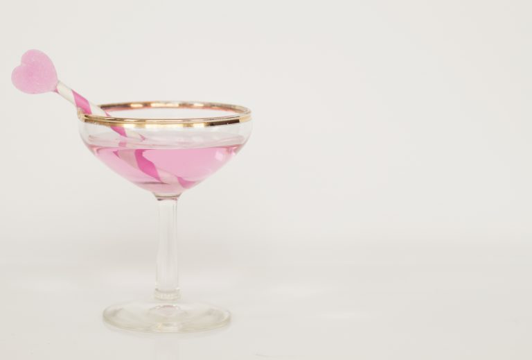 A pink alcoholic drink in a glass with a gold rim, it has a pink striped straw with a heart on top of it