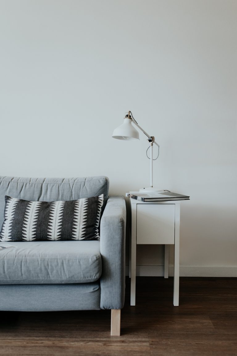 A light grey sofa up against a white wall. next to the sofa is a small white table with a white lamp and a book on it.