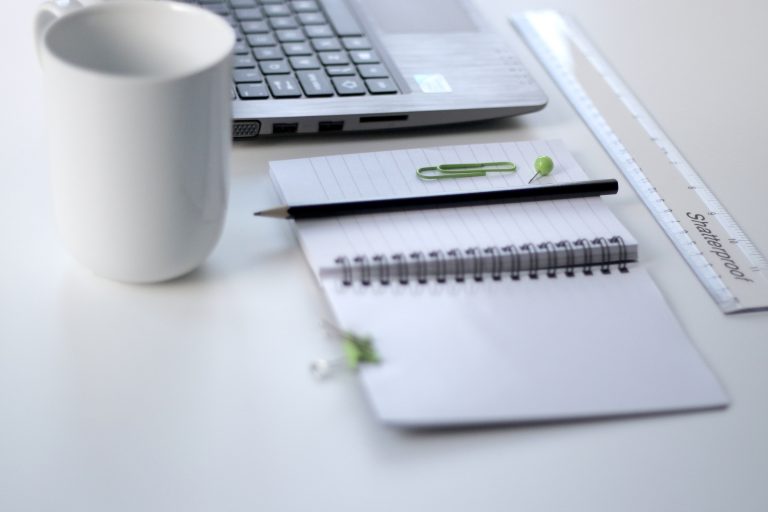 a white mug, notebook, pencil and computer sat on a white desk