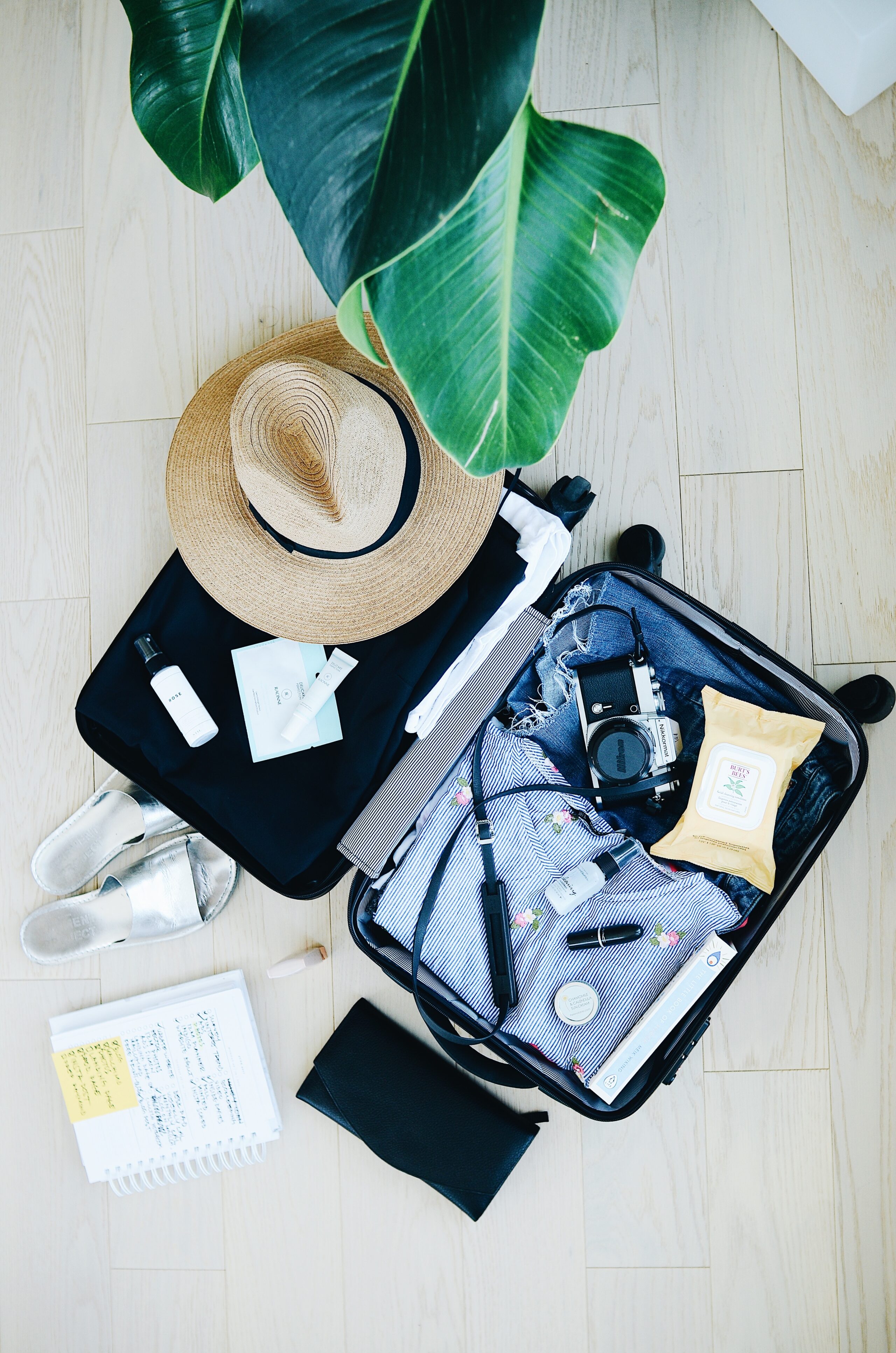 an open suitcase containing clothes and a hat, next to it is a note book, camera and passport