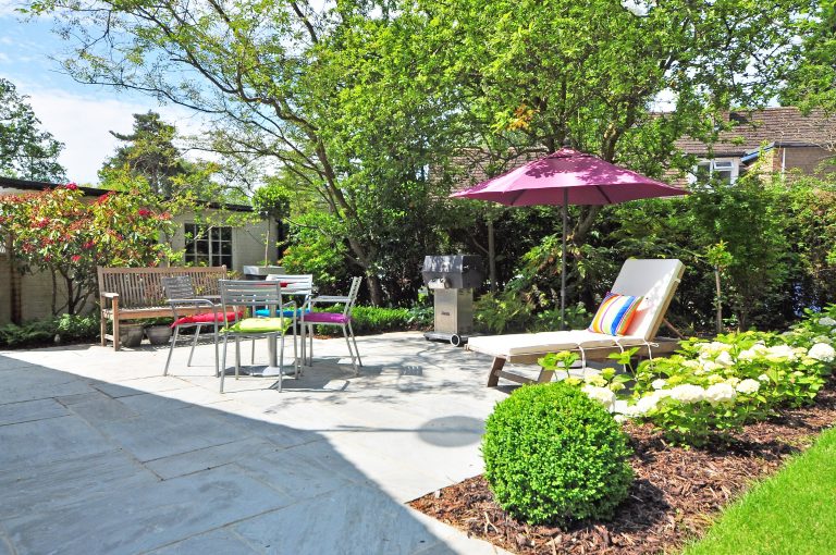 A beige garden chaise long set up under a pink parasol in a sunny garden