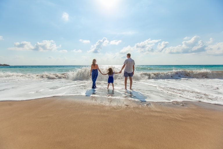 A family of three—two adults and a child—hold hands on a sandy beach, enjoying the waves and bright sun, their peace of mind supported by reliable medical insurance under the open blue sky.
