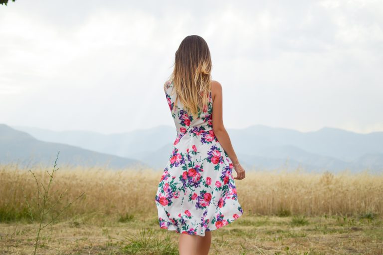A woman with long hair stands in a field, facing away, wearing one of the latest floral dresses. Mountains and a cloudy sky create a stunning backdrop.