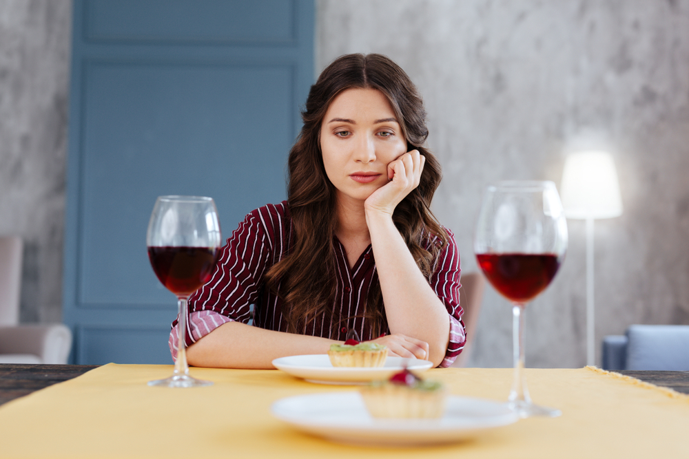 A woman sat at a table with two meals and two half full wine glasses in front of her. She looks fed up as she has her chin rested on her hand