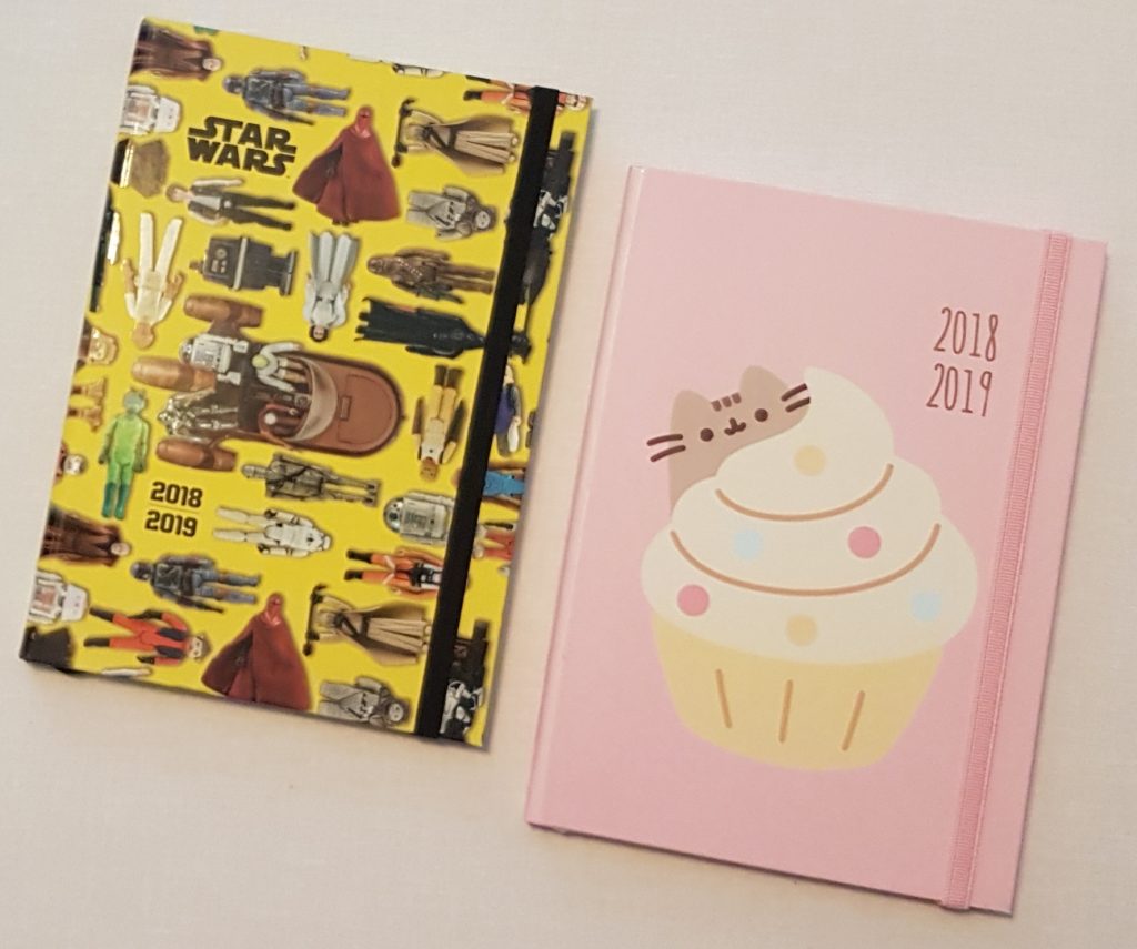 Two planners for back to school 2018–2019: one with a yellow Star Wars design featuring characters and vehicles, the other pink with a cupcake topped with a cat face illustration. Both have elastic bands to keep them closed.
