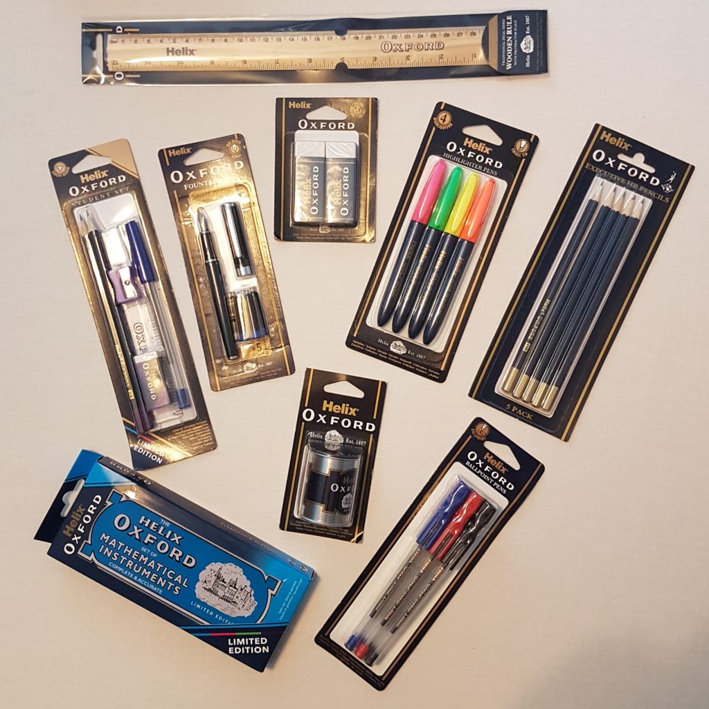 Assorted Helix Oxford stationery items on a light surface, perfect for back to school 2018, including a ruler, pencils, pens, highlighters, rubbers, a sharpener, and a blue mathematical instruments set in black and gold packaging.