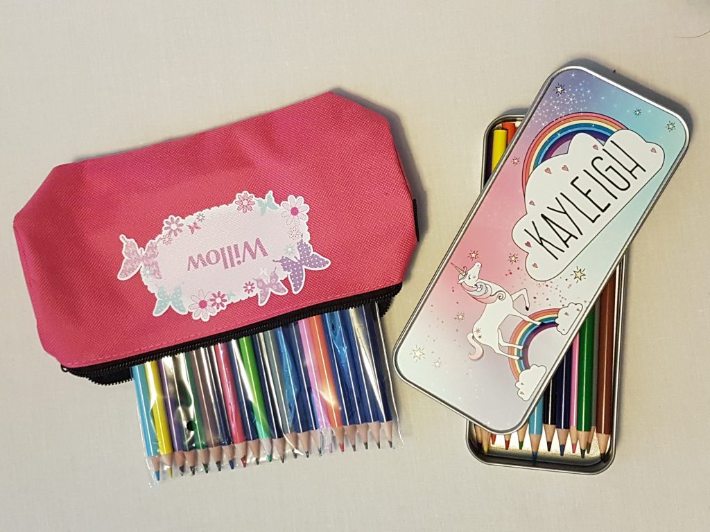 Two sets of coloured pencils—one in a metal unicorn tin labelled “Ayleigh”, and another in a pink fabric pouch with butterfly designs labelled “Willow”—perfect for back to school 2018, both displayed on a light surface.