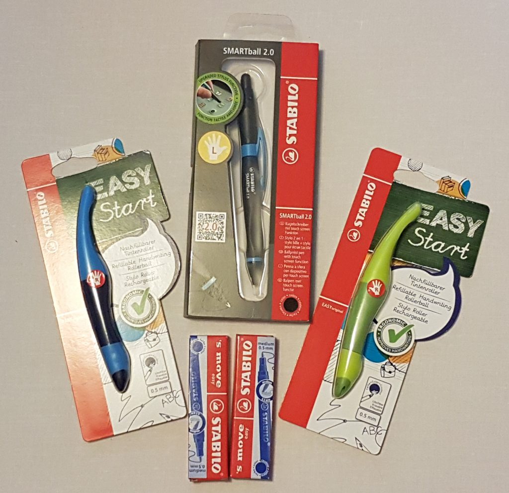 Perfect for back to school 2018, three Stabilo pens in packaging and three boxes of Stabilo ink cartridges are arranged on a light surface. The set includes two EASY Start models (blue and green) and one SMARTball 2.0 (grey and red).