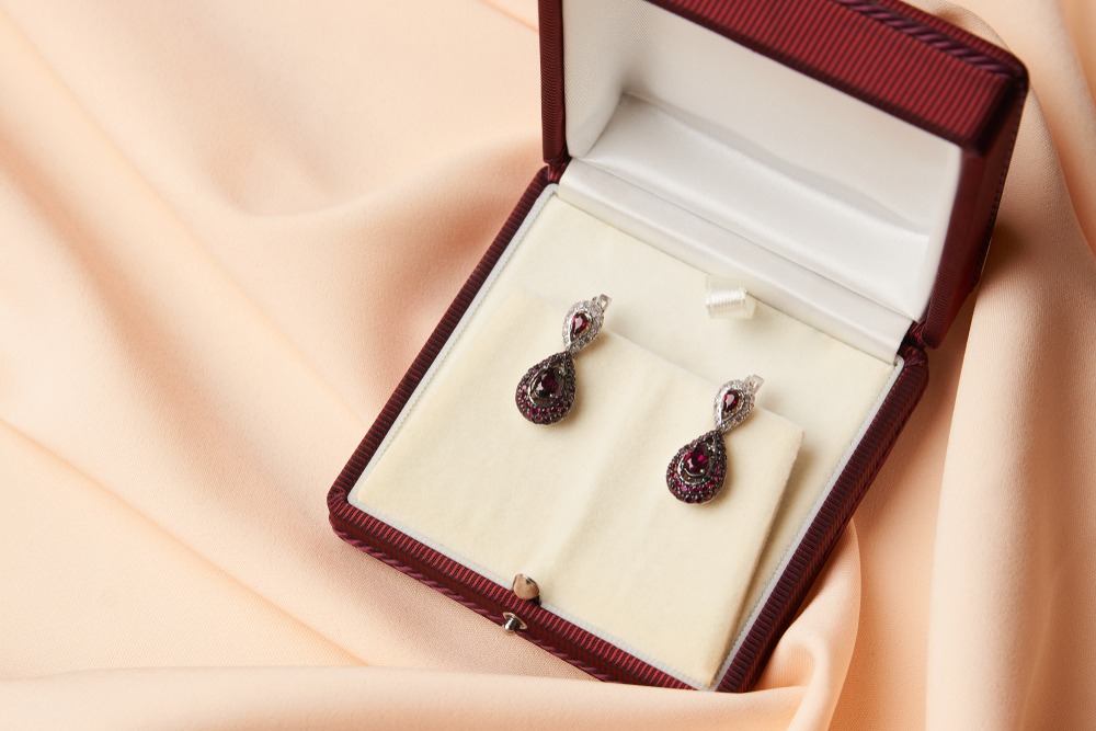 A pair of elegant, teardrop-shaped earrings with red gemstones and clear accents, displayed in an open red jewellery box on a soft, peach-coloured fabric background—perfect for those seeking luxury gifts.