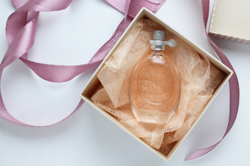 A clear perfume bottle with peach-coloured liquid rests in a cream gift box filled with peach tissue—an elegant choice for luxury gifts, complemented by a pink satin ribbon draped on the white surface.