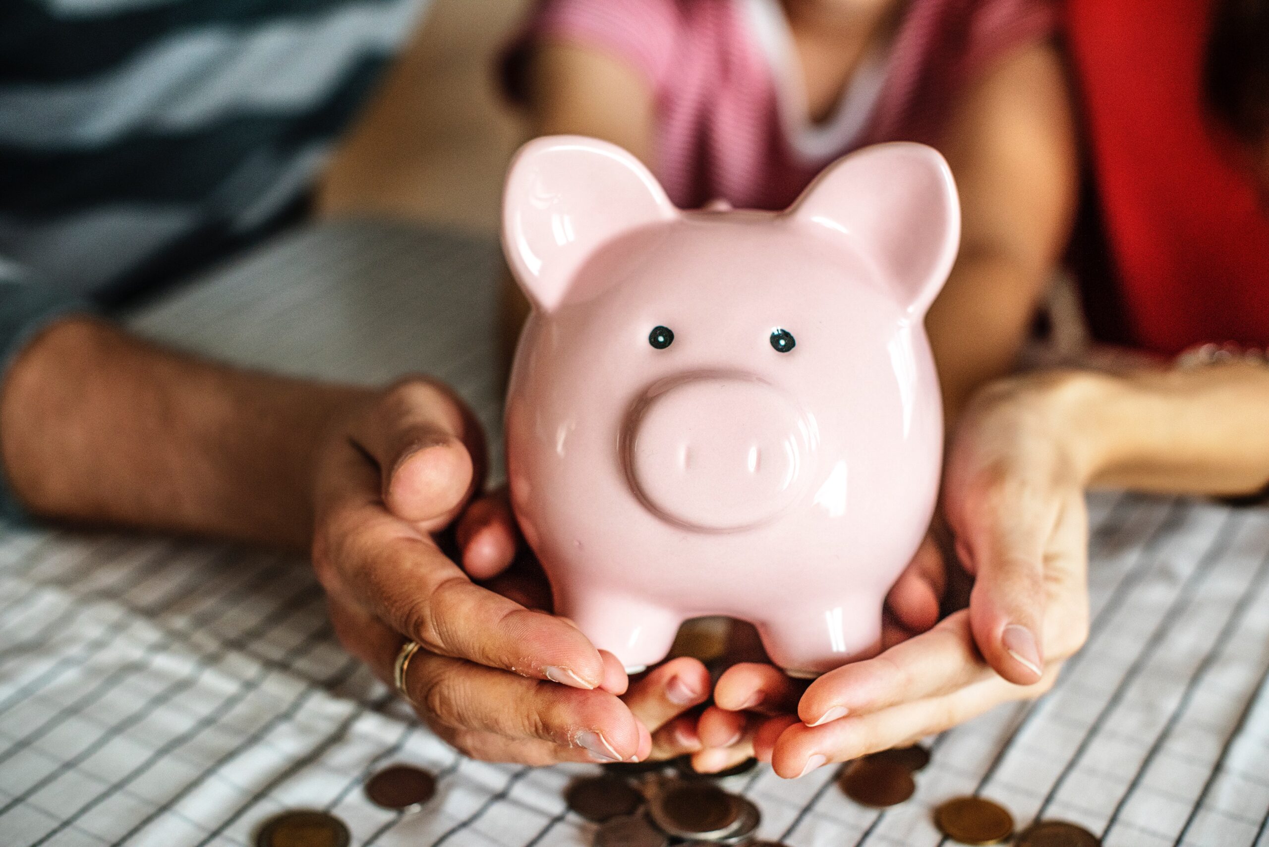 two pairs of hands holding a pink piggy bank with lots of money around it