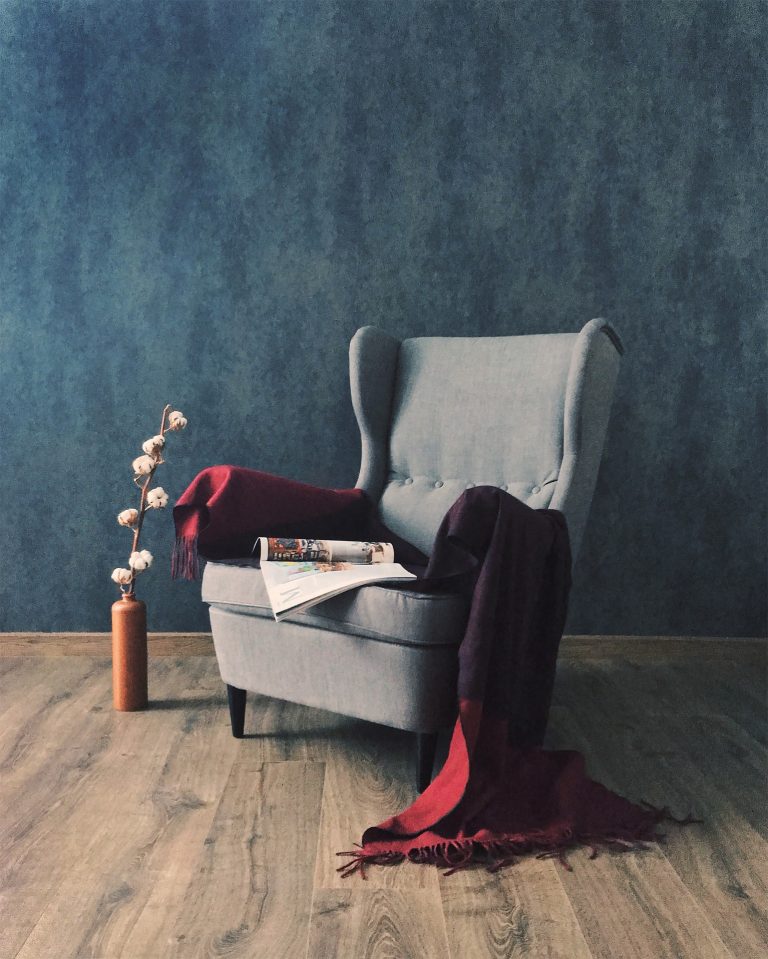 A grey highbacked armchair on a wooden floor with a blue wallpapered wall in the background