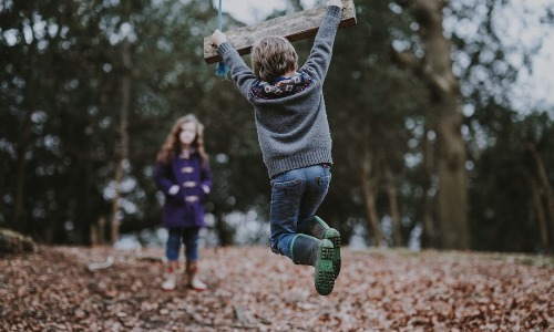 A child in jeans and boots swings on a wooden swing outdoors, whilst another child in a purple coat stands in the background among trees and fallen leaves.
