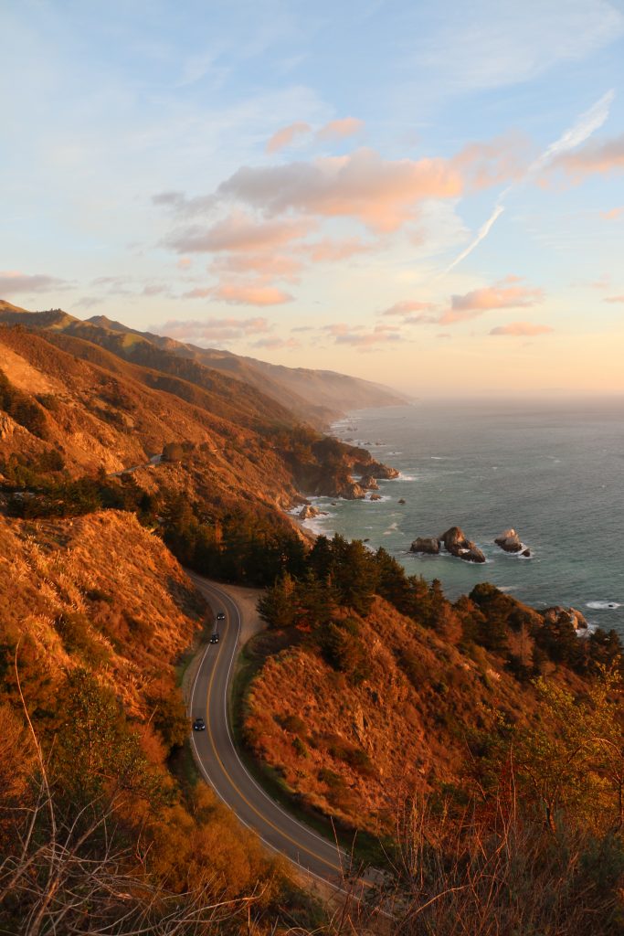 A winding California coastal road curves along rugged cliffs beside the ocean at sunset, with cars driving, waves crashing, and hills bathed in golden light under a partly cloudy sky.