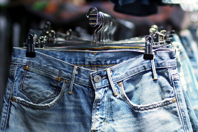 A close-up of light blue denim jeans for the autumn, with visible pockets and a button, hanging on a clothes rail with black clips in a shop. Other pairs of jeans are blurred in the background.