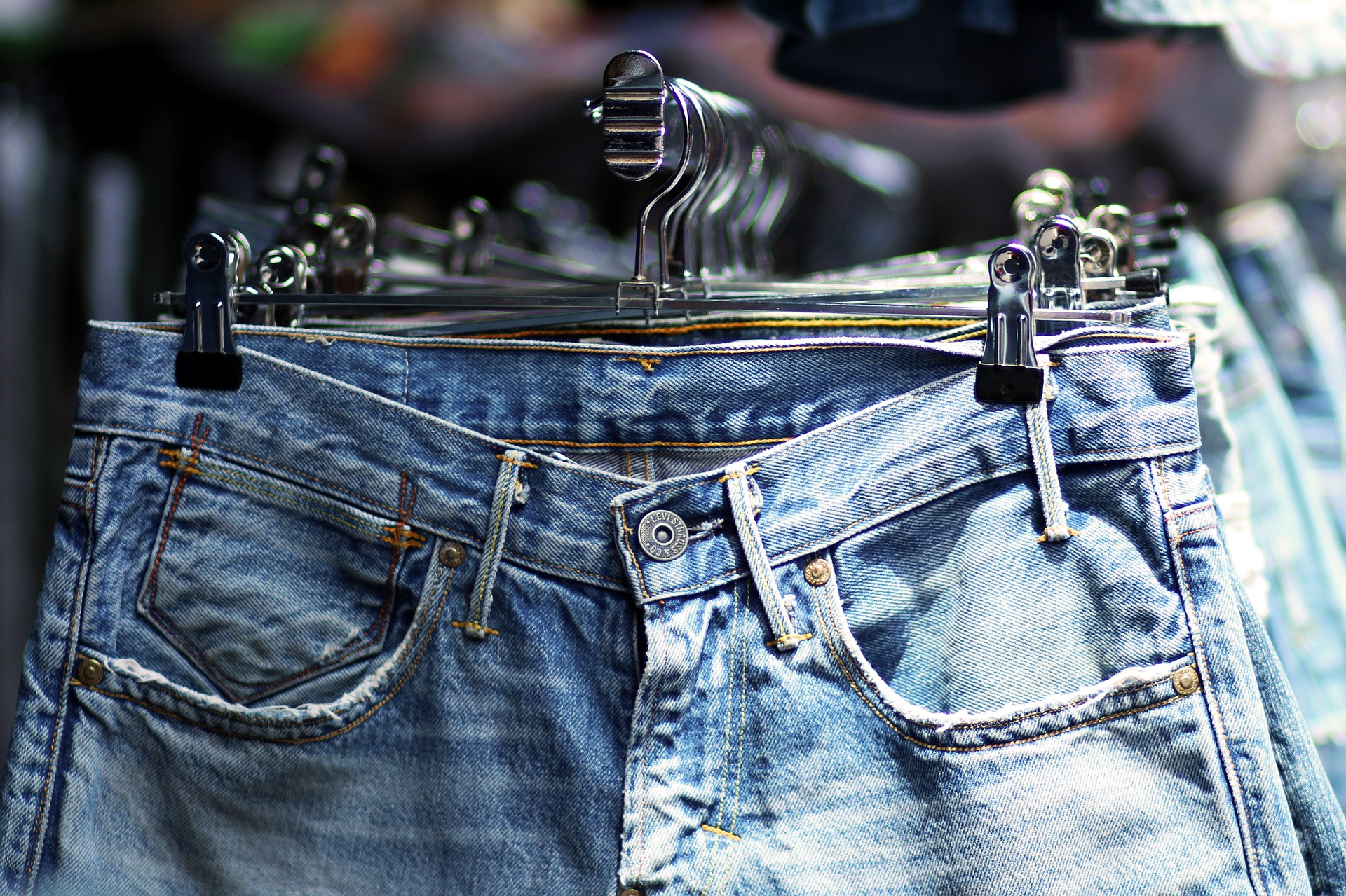 A close-up of light blue denim jeans for the autumn, with visible pockets and a button, hanging on a clothes rail with black clips in a shop. Other pairs of jeans are blurred in the background.