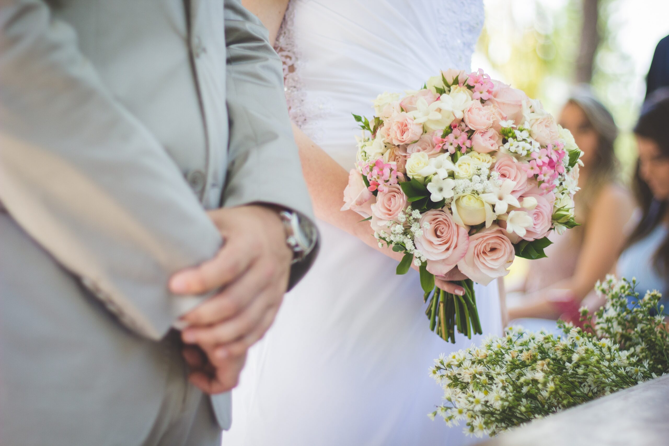 bride and groom stood together, bride is dressed in white and holding a bouquet of pink flowers, groom is wearing light grey